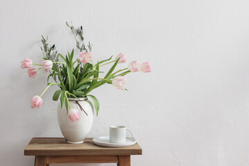 Romantic Easter, spring still life scene. Cup of coffee and floral bouquet in white ceramic vase on wooden bench. Feminine styled photo. Pink tulips flowers, olive tree branches on table. Copy space.