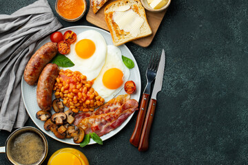 Full English breakfast on a plate with fried eggs, sausages, bacon, beans, toasts and coffee on dark stone background. With copy space. Top view