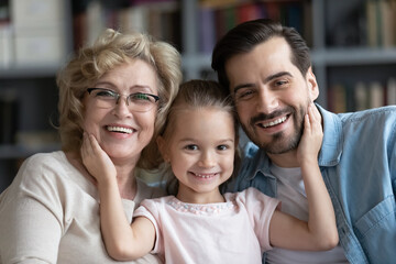Head shot portrait of happy adorable little preschool 7s kid girl cuddling affectionate middle aged older granny and loving young father, posing for photo together at home, family relations concept.