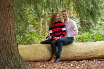 Naklejka na ściany i meble Beautiful enamored young couple sitting on a tree trunk in the forest. Facial expression is in love. In half body, man sits behind the woman