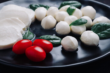 White small mozzarella cheese balls, spinach leaves and tomatoes on black plate.