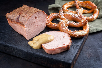 Traditioneller ofenfrischer bayrischer Leberkäse aufgeschnitten und am Stück serviert mit Brezen und scharfen Senf als close-up auf einem alten rustikalen Holz Board