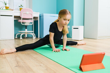 Primary school age sportive gymnast girl in black leotard stay in twine pose and look at tablet screen during online training at home