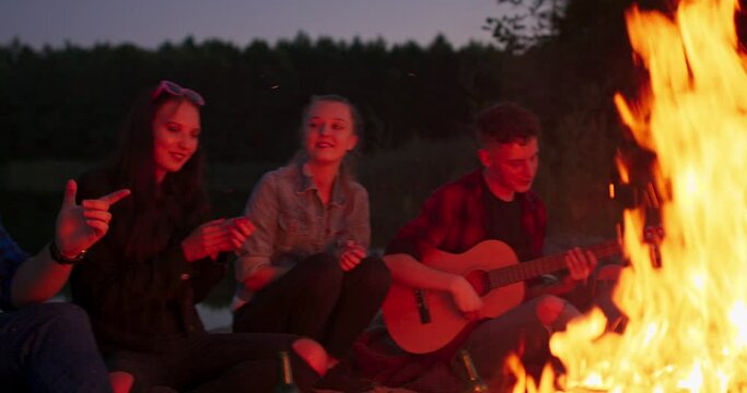 Handsome teenage boy is playing the guitar while her friends are singing funny songs and laughing and clapping hands. Music, nature and fun concept.