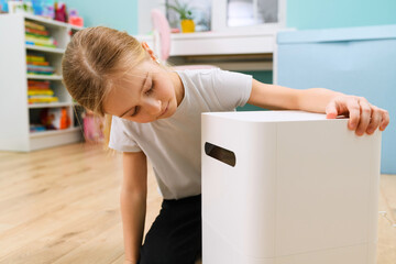 Fototapeta na wymiar Small girl sitting at room floor and looking at big air humidifier device close up for comfort home staying