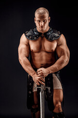 Fototapeta na wymiar Severe barbarian in leather costume with sword. Portrait of balded muscular gladiator. Studio shot. Black background.