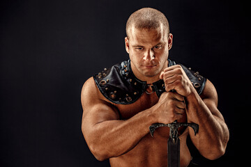 Severe barbarian in leather costume with sword. Portrait of balded muscular gladiator. Studio shot. Black background.