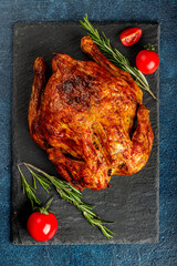 Top view or flatlay concept with grilled, roasted whole chicken on a black stone plate with rosmary and cherry tomato