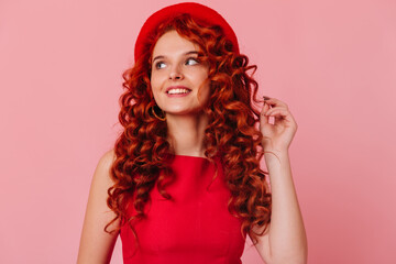 Charming red-haired girl with smile looks away. Woman in hat and red top touches hair
