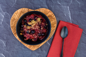 Serving of fresh baked blueberry strawberry crisp in a black bowl on a heart shaped wood board
