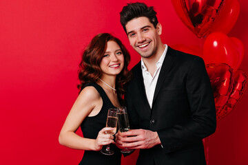 Studio shot of married couple isolated on red. Laughing young people posing with wineglasses at party.