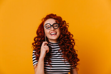 Positive woman in black and white top laughs sincerely, holding toy glasses. Portrait of curly redhead girl in orange studio