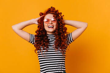 Energetic girl in striped T-shirt and pink sunglasses ruffles her curly red hair and smiles