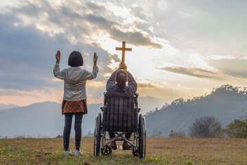 Silhouette of disabled woman sitting on her wheelchair and hold the crucifix in hands with child...