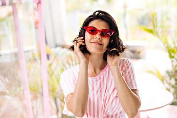 Front view of stunning european girl in striped t-shirt. Smiling woman in pink sunglasses touching hair on blur background.