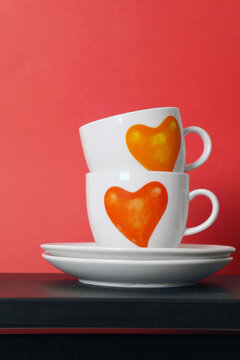 Two white tea cups with painted red hearts on red background, selective focus,copy space