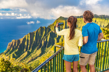 Hawaii Kauai island travel tourists couple taking phone picture of Na Pali Coast mountain range at...