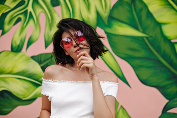 Naklejka premium Front view of interested tattooed girl in sunglasses. Blissful young woman standing in front of green graffiti.