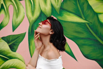 Naklejka premium Pleased tattooed girl posing in front of graffiti. Outdoor shot of glad young woman in pink sunglasses.