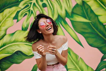 Excited lady in pink glasses laughing at camera. Outdoor shot of charming girl posing near graffiti.