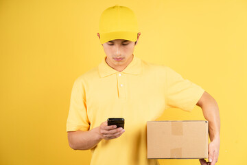 Portrait of delivery male holding cargo box and using phone on yellow background