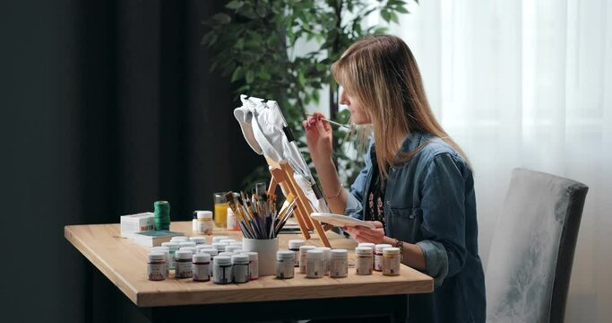 Focused young woman sitting at table with various art tools and painting on casual grey t-shirt. Female artist creating unique design on clothes.