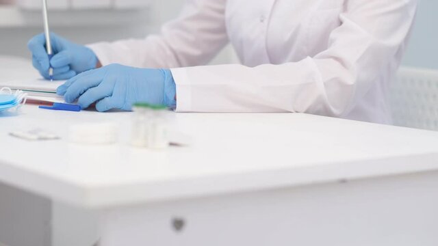 4k Unrecognizable woman doctor surgeon in white uniform is writing diagnosis and examination results. Hands in blue gloves close up. Healthcare and medicine