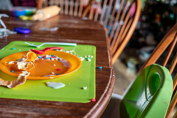 Mess left at the kitchen table after a toddler finished.  Plastic fork, cup and plate strewn with drips and crumbs.