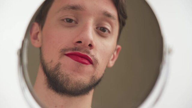 One young Caucasian bearded brunette handsome male with painted lips look at himself in round mirror, flirt, blow kiss on white background. Face of beauty transvestite or gay man admires close up.