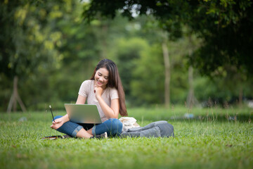 Confident smiling young Asian woman students reading and thinking doing homework with computer laptop on lawn in park. Concept of Education, Campus, teenage.