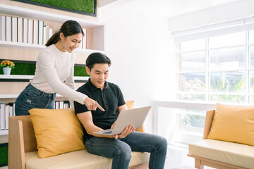 Asian male sitting and female standing back of him while working, pointing on the laptop together. Businessman working on laptop at home. Good opportunity comes with a surprise. Focus on a man.