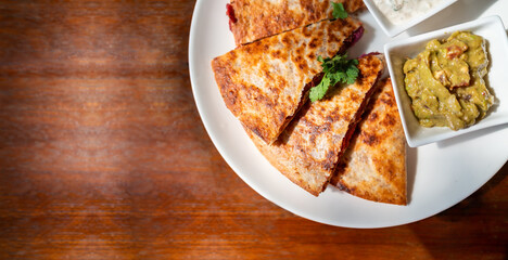 Crop image top view of Vegan Quesadilla: grilled whole-wheat tortilla filled with sweet potato and red bean, vegan quesso cheese served with guacamole salsa and Tzaziki sauce.