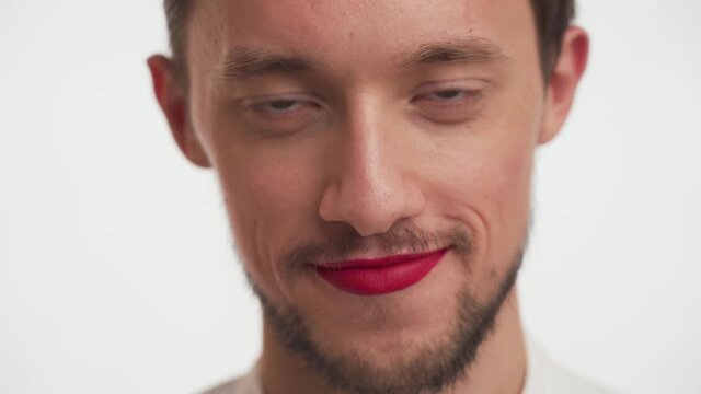 Close-up portrait of bearded male with painted lips by lipstick. Attractive guy with moustache looking at camera and down, flirting, shy on white background. Concept of handsome effeminate young man.