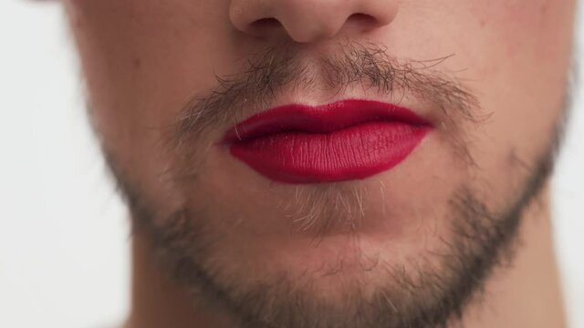 One young metrosexual or gay man with dark brown beard, moustache wear makeup – red lipstick on lips, talk, smile, blow kiss on white background indoor. Close up face of handsome guy like transvestite