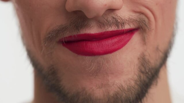 Hint of sex. One sexy Caucasian young man with brown beard, moustache, red lipstick on lips showing oral symbol with tongue and mouth. Cute male make forbidden gesture on white background close up.