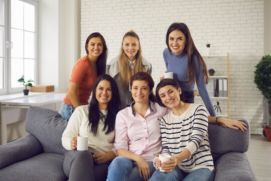 Group of young women sitting on sofa at home, drinking tea, smiling and looking at camera. Happy friends and coworkers enjoying Casual Friday at work and spending coffee break on modern office couch - Powered by Adobe