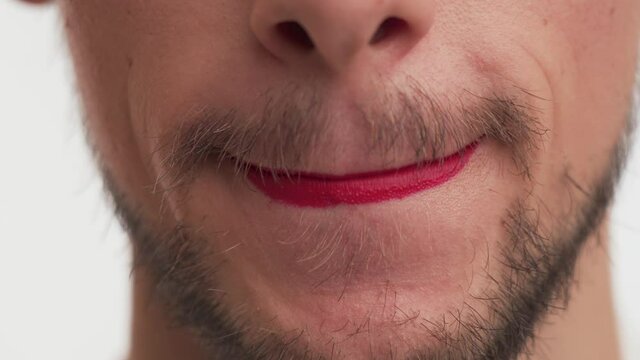 One young metrosexual or gay man with dark brown beard, moustache wear makeup – red lipstick on lips, speak some phrase, on white background indoor. Close-up mouth of handsome guy like transvestite.