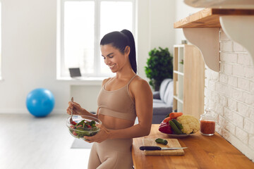 Slender fitness woman in sportswear standing at home in the kitchen with a bowl of fresh salad. Happy woman enjoys healthy food after workout. Fitness, dieting and healthy lifestyle concept. - obrazy, fototapety, plakaty