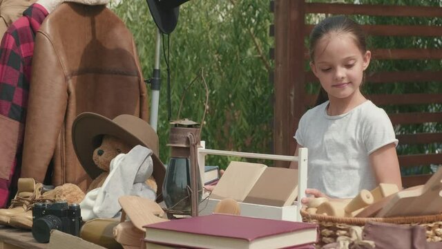  Handheld tilt up of little girl organizing second hand items on table in front yard on summer day and preparing for garage sale, then looking at camera and smiling