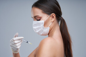 Portrait of beautiful woman holding vaccine in mask on gray background