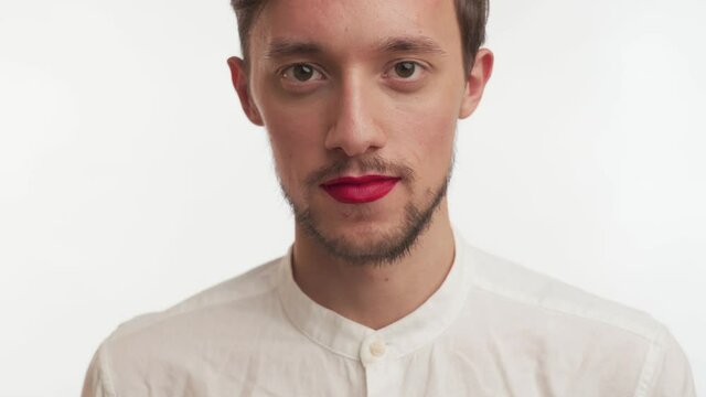 Serious beautiful man with brown eyes, beard, painted lips by red lipstick wear shirt, stare at camera, smile, laugh isolated on white wall background. Portrait of single transvestite happy model guy 