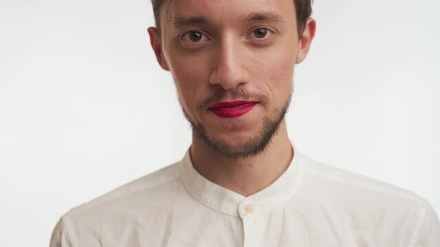 Young Caucasian man with brown beard chews food, swallows, blow kiss, smile isolated on white background. Handsome guy with brown eyes, painted lips by red lipstick stand, stare at camera close up.