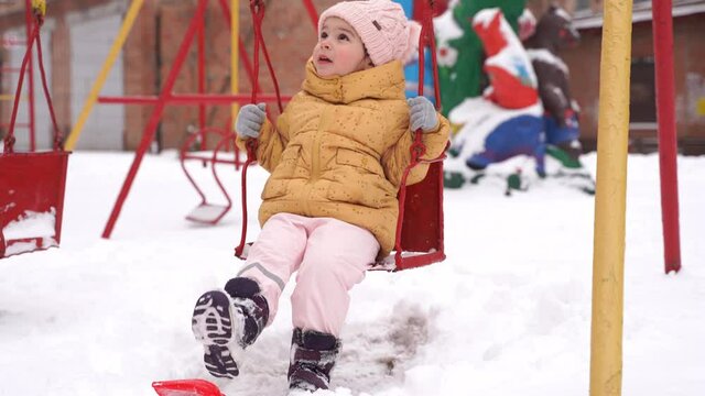 Toddler girl playing on playground in winter. Snowy day on swing. Kids winter activity with snow. Cold weather snowsuit for child.