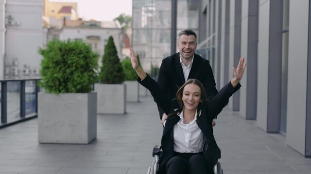 Cheerful man pushing wheelchair with his disabled coworker and smiling near office building. Male and female happy businesspeople having fun together outdoors. Concept of support.