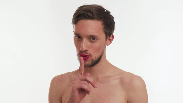 Single young cute shy half naked Caucasian brunette man with brown beard puts finger to painted lips by red lipstick, show gesture shh silence, blow sweet kiss isolated on white background close up.