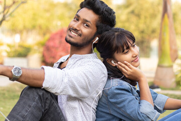 Obraz premium Young teenage friends enjoying listening to favorite song or music by Sitting back to back at college - Concept of couple relaxing, picnic in park,Love, dating, romance and modern millennial lifestyle