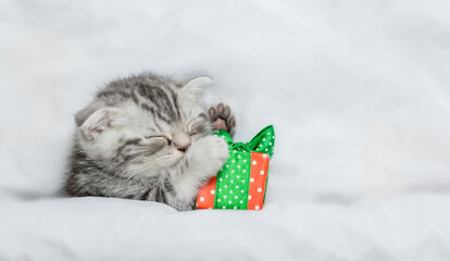 Cute tabby kitten sleeps under white blanket and hugs gift box. Top down view. Empty space for text