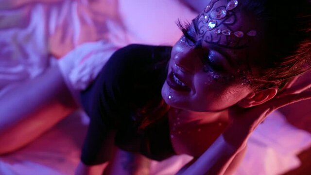 A woman with a beautiful unusual makeup, a pattern and with rhinestones on her face poses in front of the camera in the studio sitting on the bed.