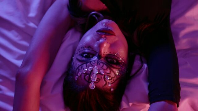 Face a model with beautiful makeup, a pattern and with rhinestones on her face poses beautifully in front of the camera in the studio lying on the bed.