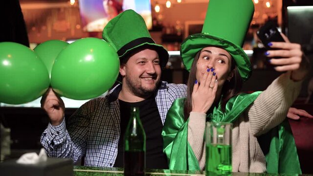 man and women leprechaun celebrating st patrick's day sitting in a bar drinking green beer and waving balls. Taking a selfie on the phone